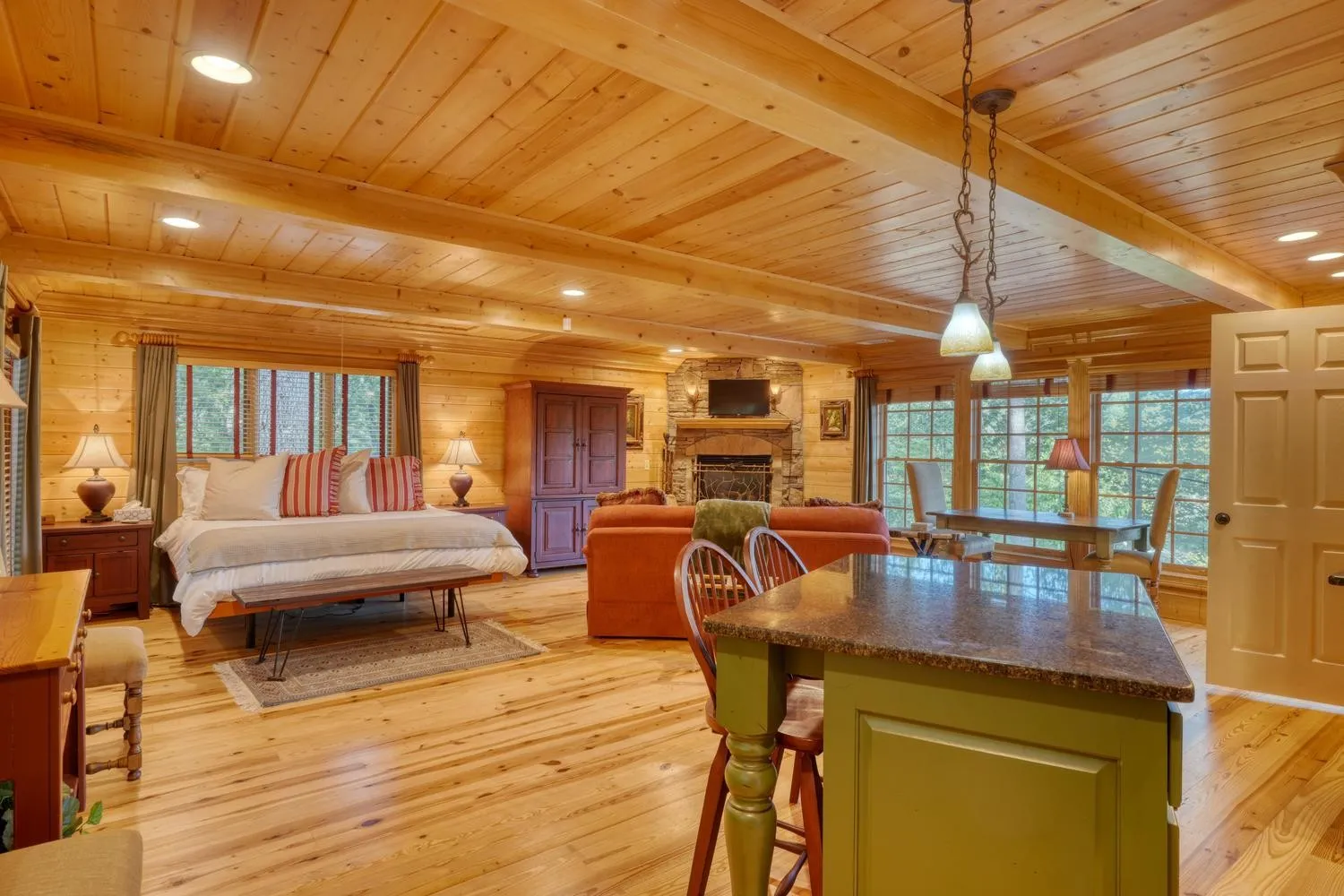 Carriage house interior with a king bed nook, an orange sofa facing a stone fireplace with a mounted TV, and a green kitchen island with barstools open to a dining table by tall windows